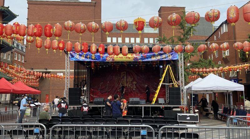 Trailer stage set up in a square in london, with Chinese lanterns hanging above it. Crews are setting up the equipment.