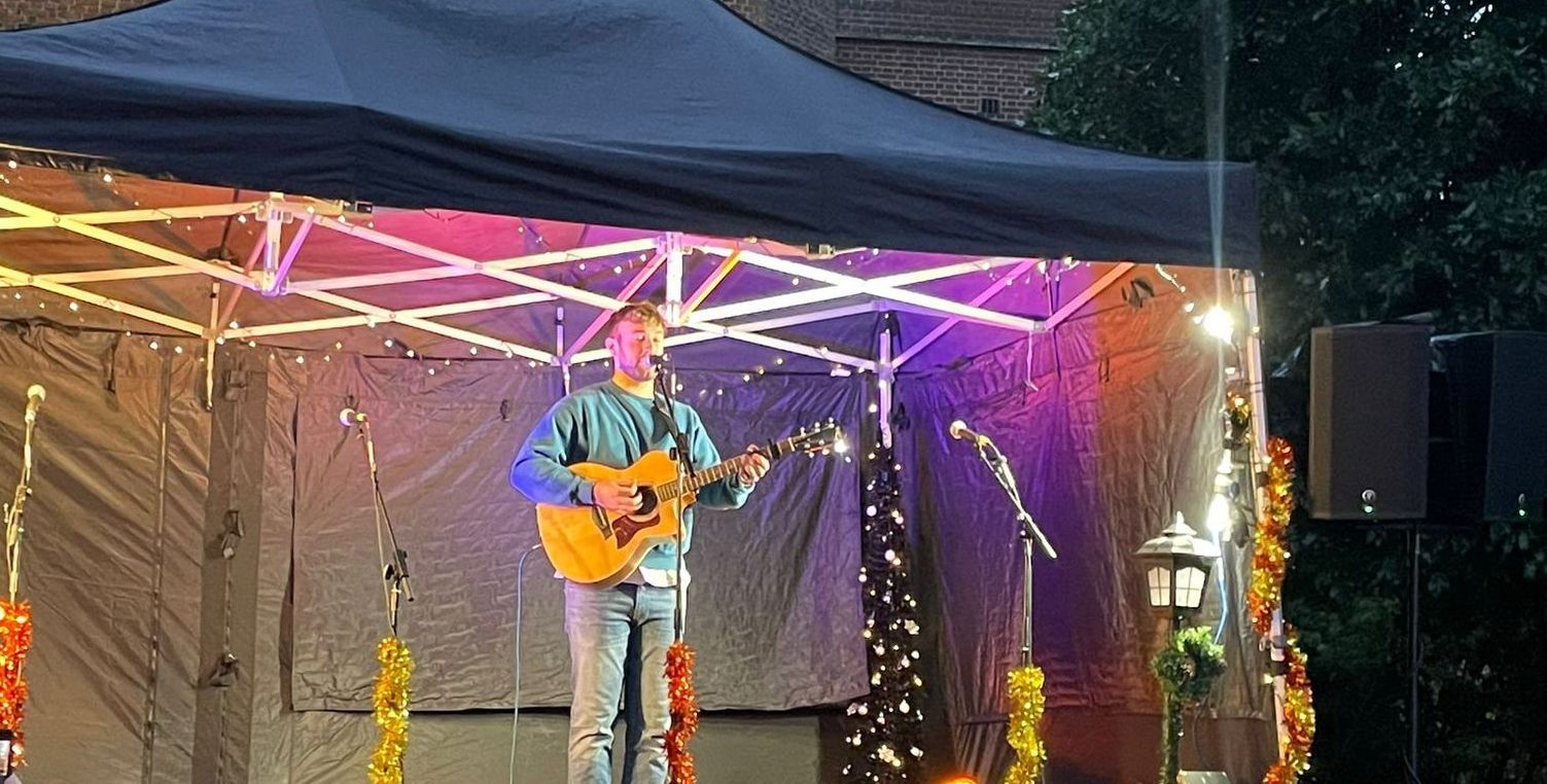 A close up of a modular stage with gazebo canopy with a solo singer with a guitar on it. The canopy is decorated with tinsel, fairy lights and a small Christmas tree in the corner.