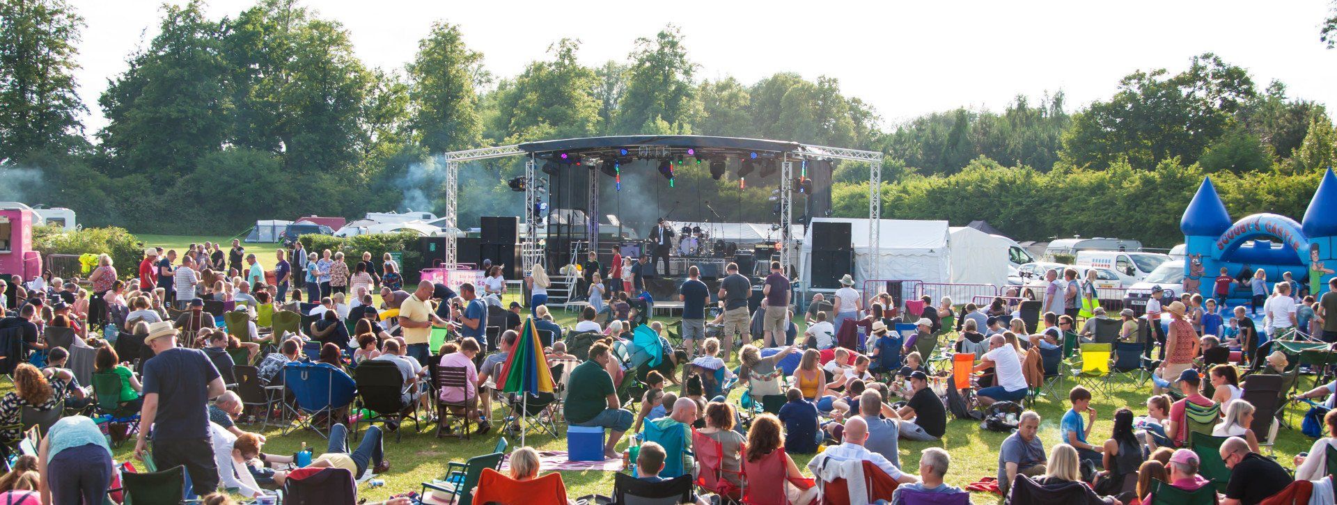 Trailer stage with PA wings and colourful lights with a band playing on it. A large audience is spread out in front of it.