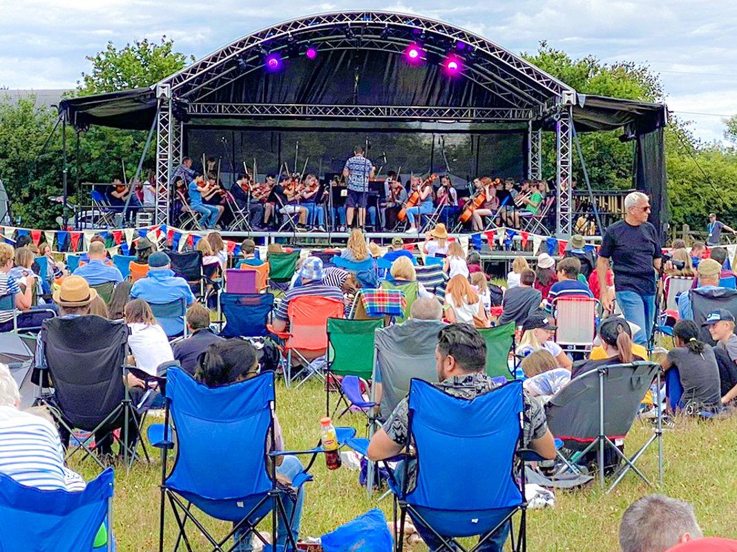 Full orchestra performing on an arc roof stage with an audience