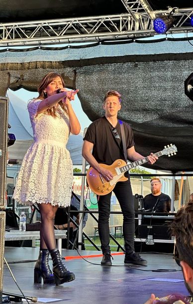 A close up shot of Natalie Imbruglia performing on an arc truss stage from Koncept Productions, with a guitarist. There are blue lights above and she wears a white dress and black books. The photo is from the foodies festival 2023