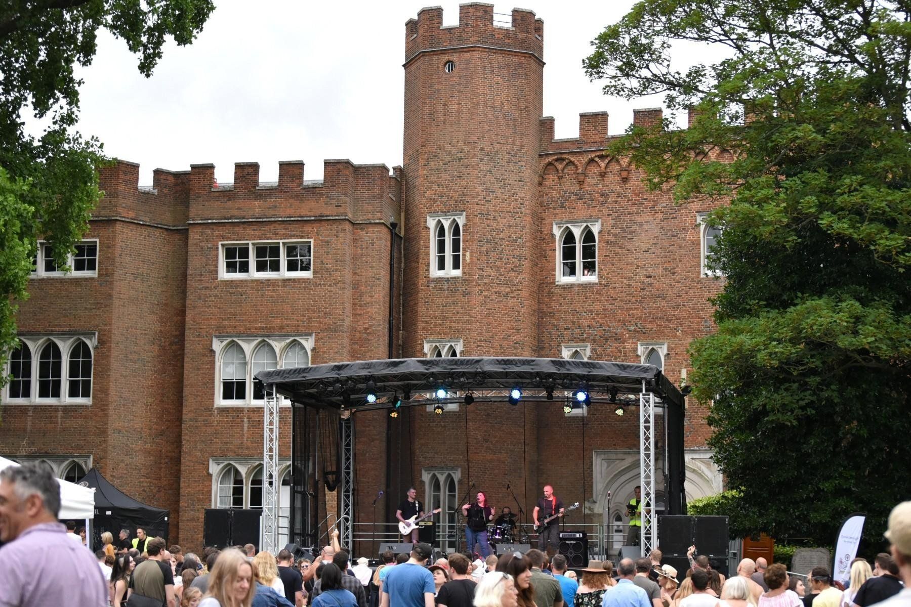 Full view of a trailer stage in front of Hertford Castle with a band performing on it and lighting hanging from the trusses