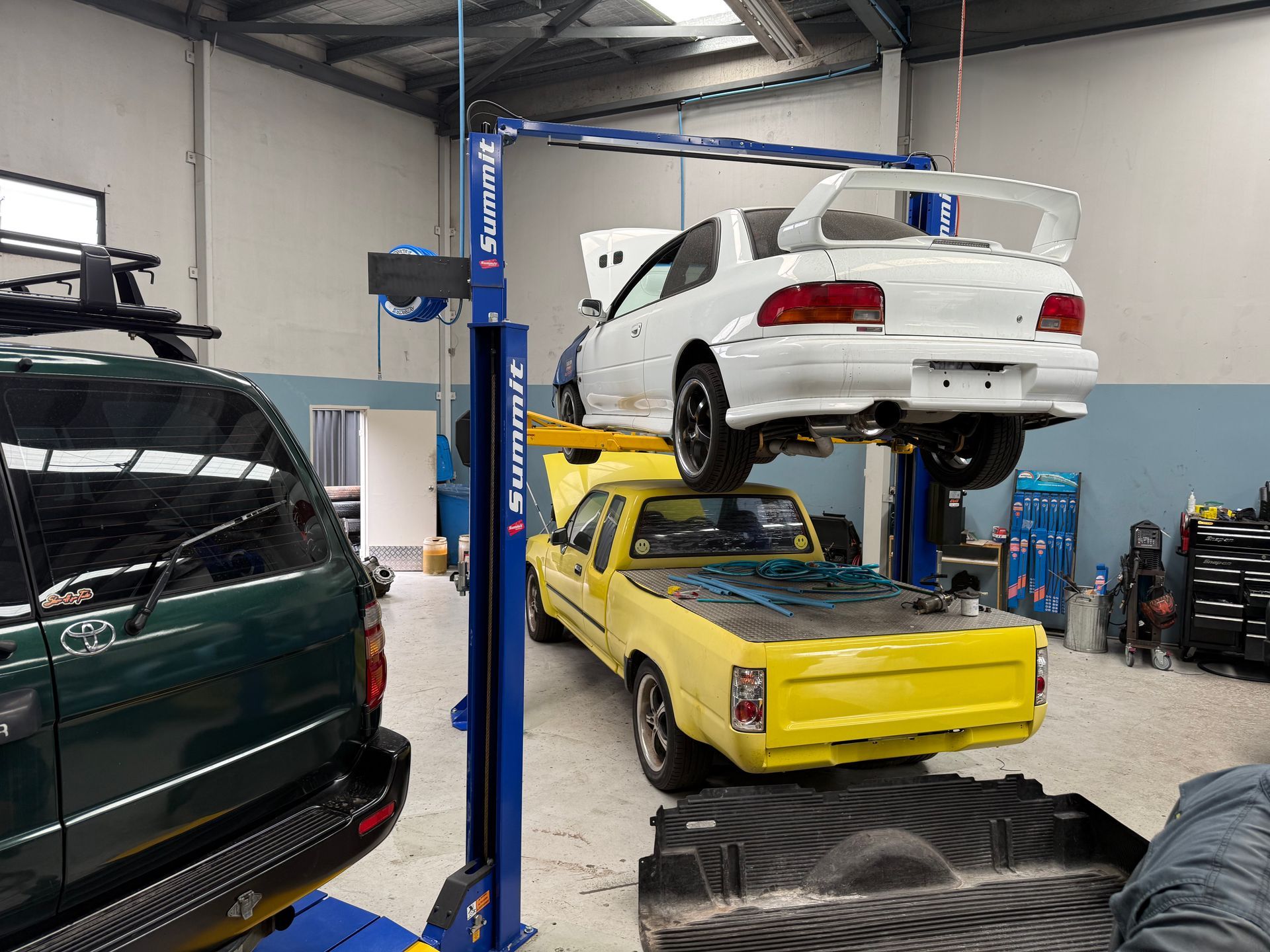White Sedan Hoisted on a Blue Mechanical Vehicle Lift Above a Pickup Truck — Mad Monkeys Mechanical in Cairns City, QLD
