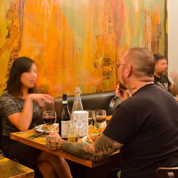 Woman and man seated at a restaurant table, conversing. Red wine, food, and colorful art backdrop.