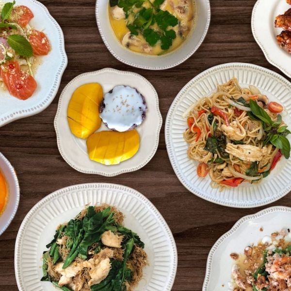 Overhead view of various Thai dishes on white plates, mango sticky rice in center, on a wooden table.