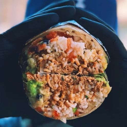 Hands holding a burrito cut in half, revealing rice, meat, avocado, and vegetables.