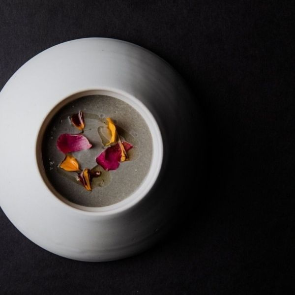 White bowl with gray soup, garnished with colorful flower petals, on a black surface.