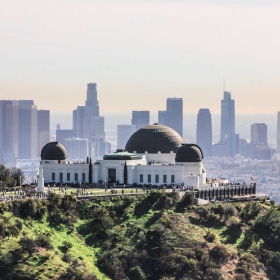 Griffith Observatory on a hill with the Los Angeles skyline in the background. White building, blue sky.