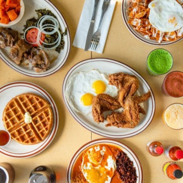 Overhead view of a breakfast spread: waffles, fried chicken, eggs, chilaquiles, sides, and drinks on a table.