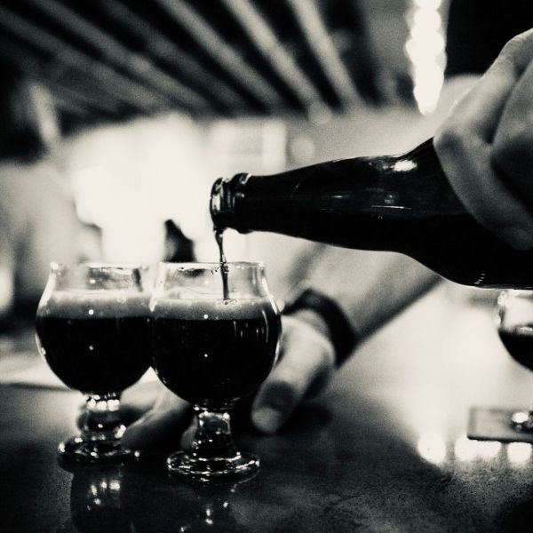 A person pours dark beer from a bottle into three tulip-shaped glasses on a bar.