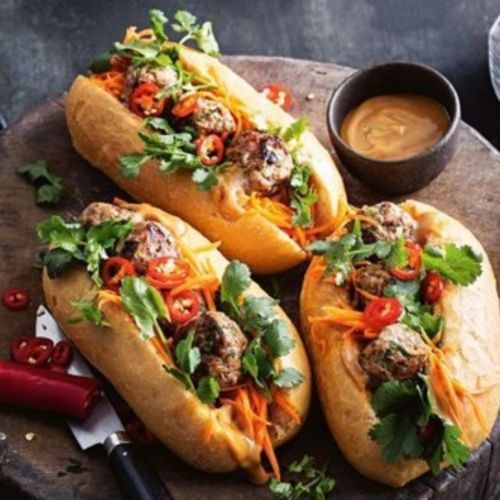 Three meatball sandwiches on a wooden board, with cilantro, carrots, and dipping sauce.