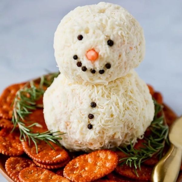 Snowman-shaped cheese ball with olive eyes, carrot nose, and peppercorn mouth, served with crackers and rosemary.