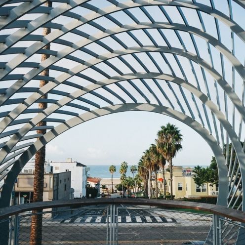 Arched metal structure frames a coastal street with palm trees, buildings, and a distant ocean view.