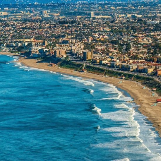 Aerial view of a coastal city with a sandy beach and blue ocean. Buildings line the shore.