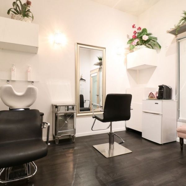Hair salon interior: black chair, mirror, shampoo bowl, white walls, and dark wood floor.