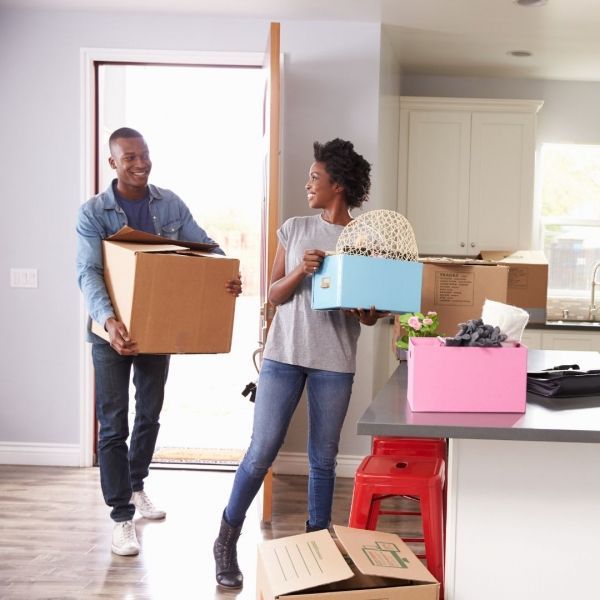 Couple carrying boxes, entering new home; man smiles carrying large box, woman with small box and decor.