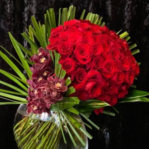 Red roses and dark orchids in a glass vase with green foliage against a black background.