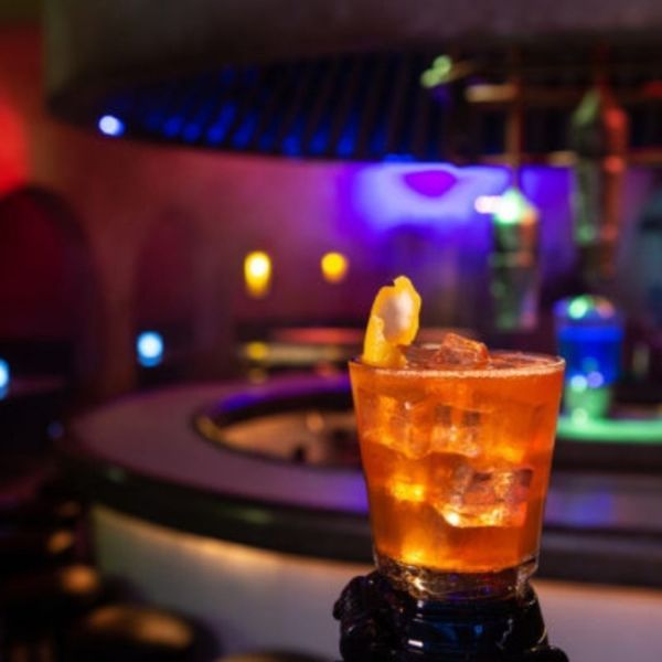 Cocktail glass with ice and a lemon twist on a bar; a blurred, dimly lit bar in the background.