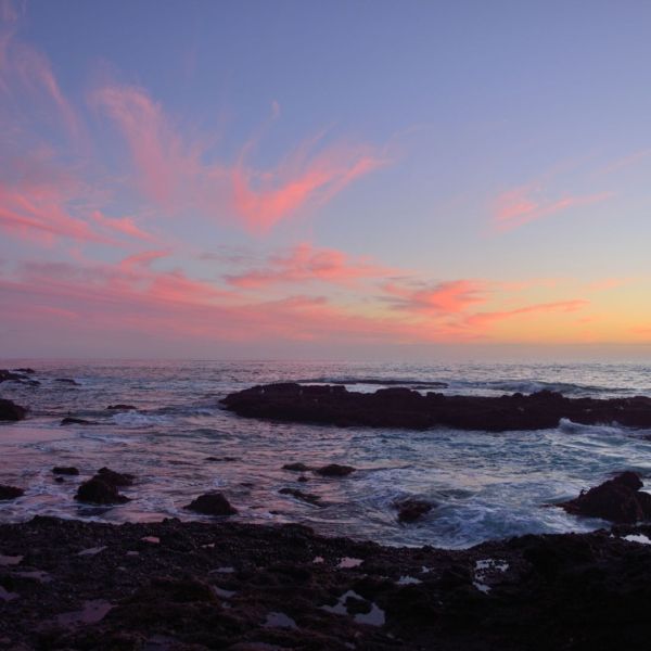 Ocean at sunset, pink and orange clouds above dark rocks and waves.