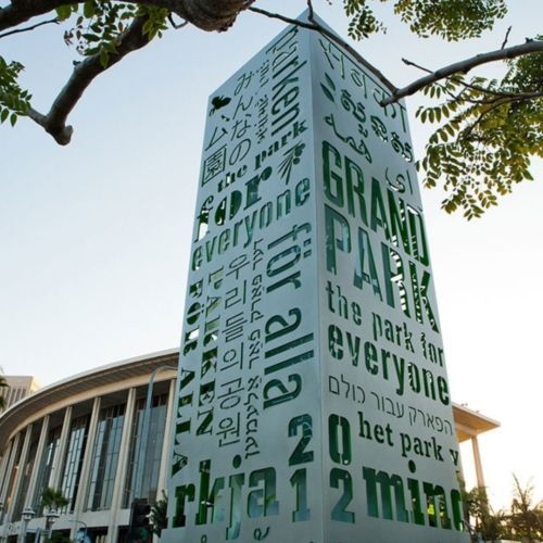 A tall monument in Grand Park, with “Grand Park” in multiple languages, with a curved building in the background.