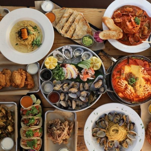 A table laden with various seafood dishes: oysters, pasta, tacos, fried food, and bowls of soup.