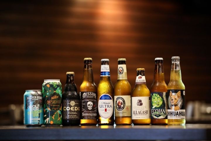 Ten various beer bottles and a can lined up on a bar with a wooden background.