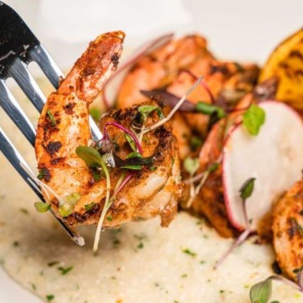 Fork lifting a grilled shrimp from a creamy grits dish, garnished with radish slices and microgreens.