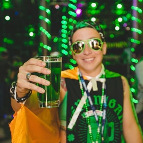 Person in green sunglasses holding a green beer, celebrating St. Patrick's Day in a festive, green-lit setting.