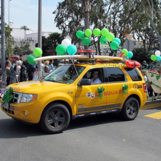 Yellow lifeguard vehicle in parade, decorated with balloons and a surfboard.