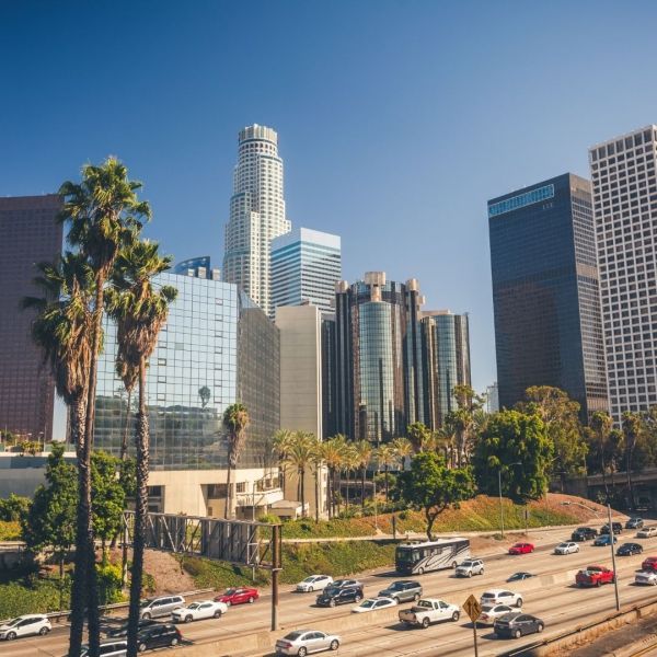 Downtown Los Angeles skyline with palm trees in the foreground, cars on a freeway below a clear blue sky.