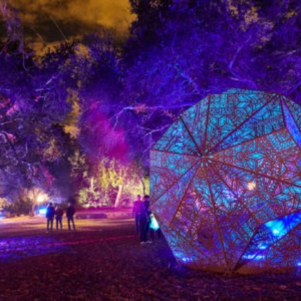 Large, illuminated geometric sculpture in park with people, under purple and blue lights.