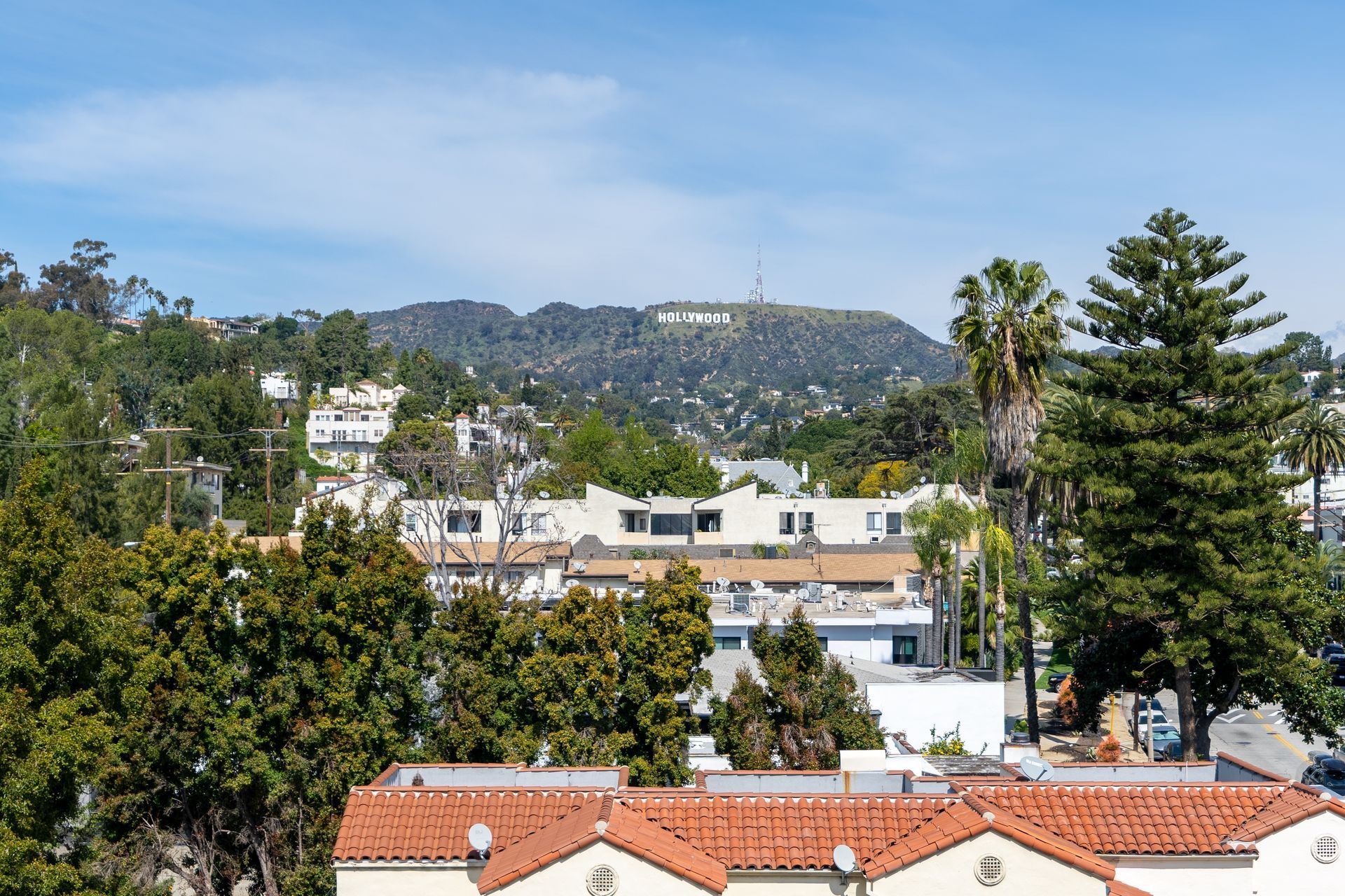 Hollywood sign visible on a hillside, above buildings and trees under a blue sky.