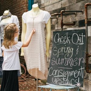 A girl touches a dress on a mannequin outside a consignment shop. A sign advertises spring/summer clothes 