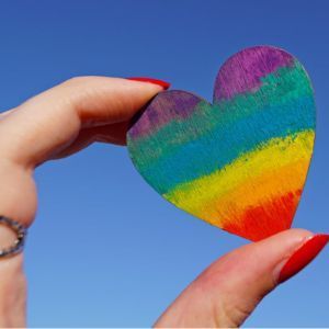 Hand holding a rainbow heart against a clear blue sky.