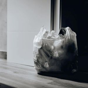 Plastic garbage bag, full of trash, sits on a wooden floor near a closed door.