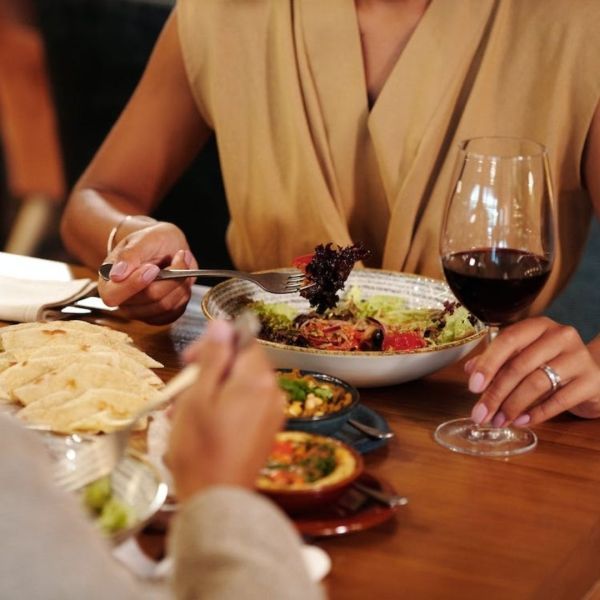 Person eating salad, holding wine glass at restaurant table with bread and small dishes.