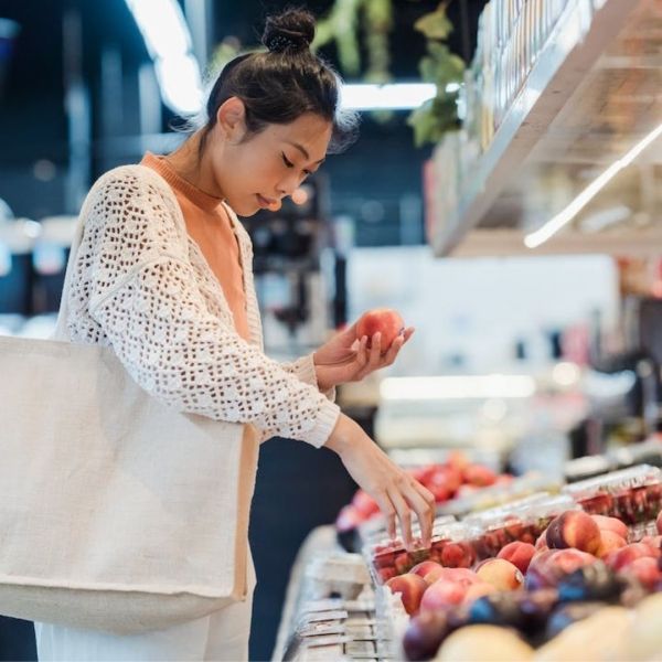 Woman selecting peaches in a grocery store, carrying a reusable bag.