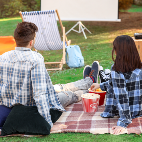 Couple watching a movie outdoors on a blanket with popcorn; screen, chairs and grass in the background.