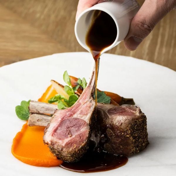 Lamb chops with sauce being poured, served with vegetables, on a white plate.
