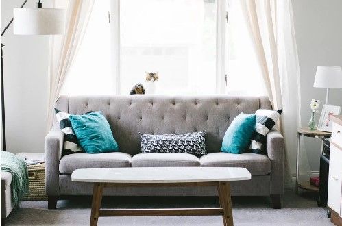Gray couch with teal pillows in a bright living room, light curtains, and a wooden coffee table.