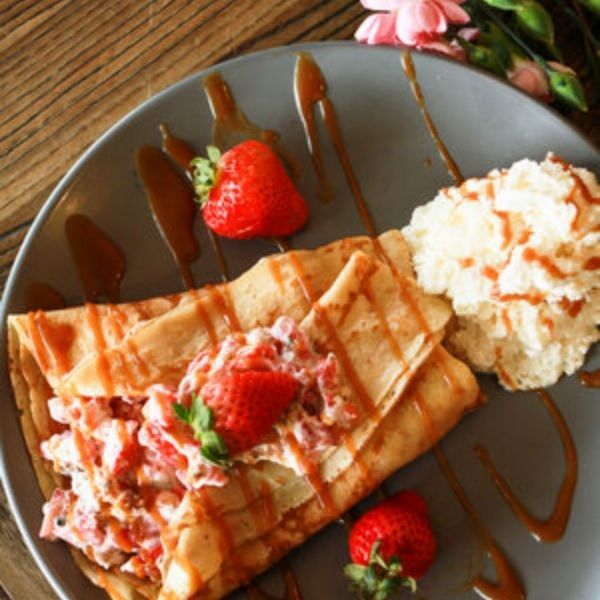 Crepe with strawberries, whipped cream, and caramel sauce on a grey plate.
