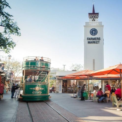 Green trolley car on tracks in front of a white clock tower at the Farmers Market. People sit and walk around.