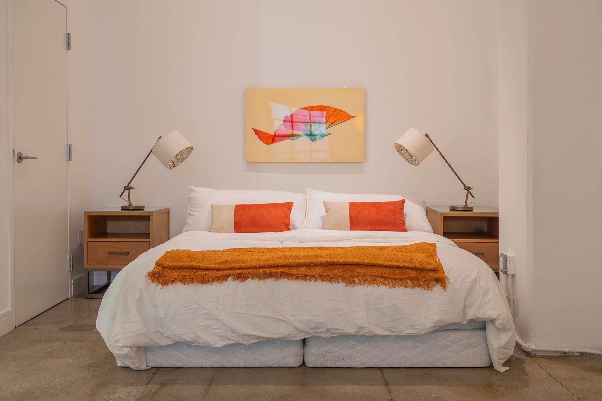 Bedroom with bed, nightstands, lamps, art, and orange throw and pillows against a white wall.