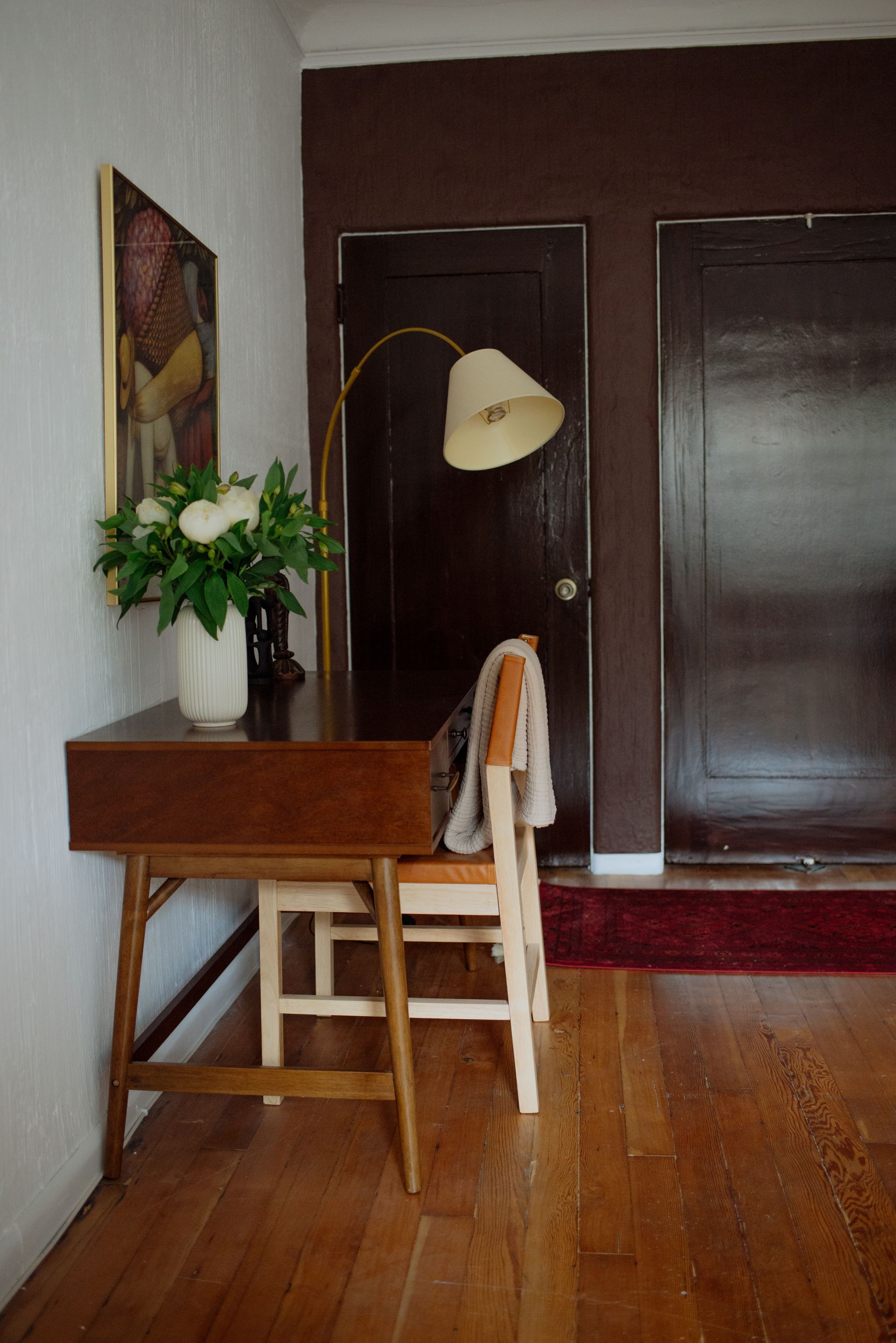 Desk and chair against a white wall with a floral arrangement, a brown door, and wood floor.
