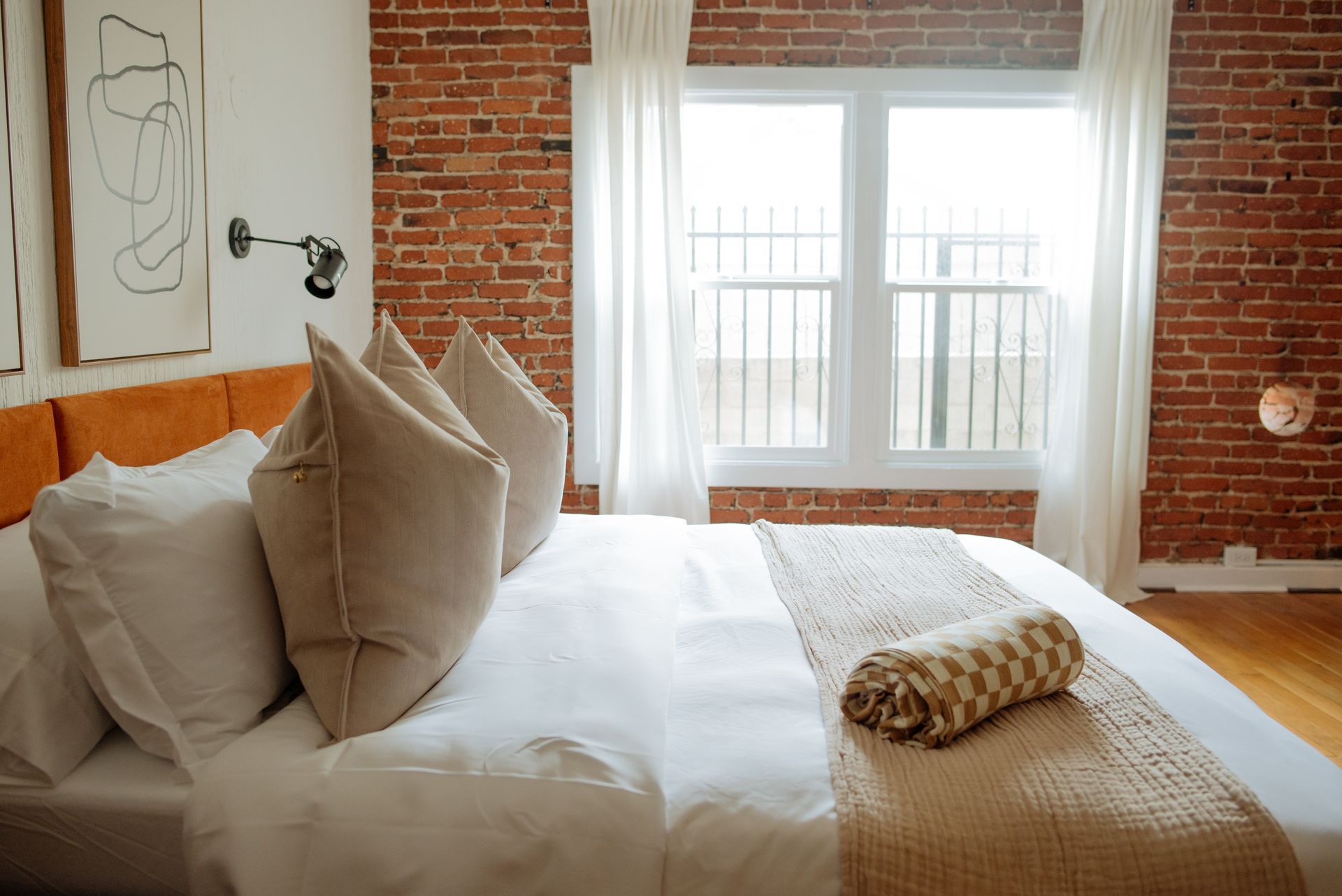 Bedroom with exposed brick walls, white bedding, and beige pillows. Orange headboard and window.