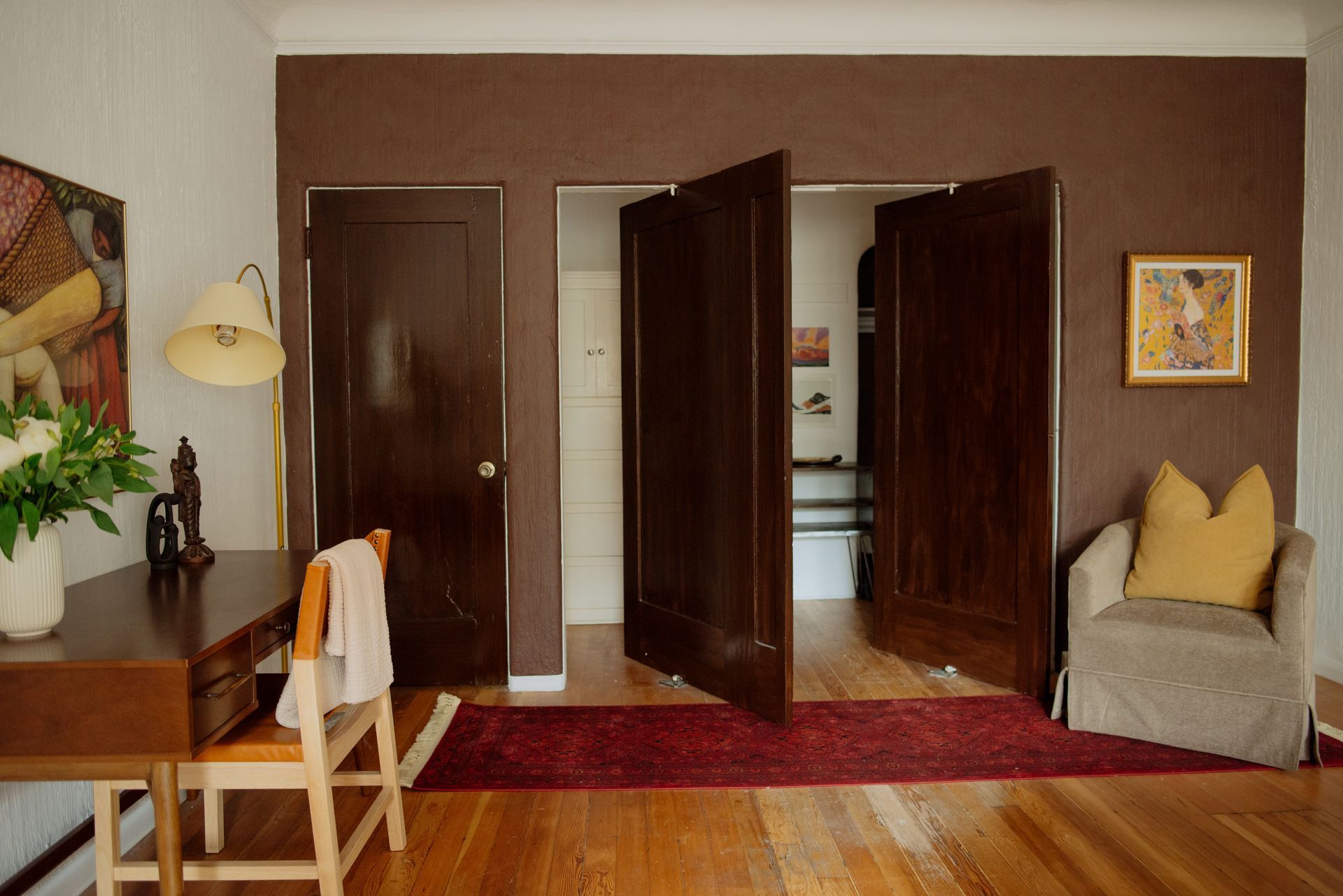 Room with three dark brown doors, one open. Brown walls, wooden floor, desk with lamp, chair, and armchair.