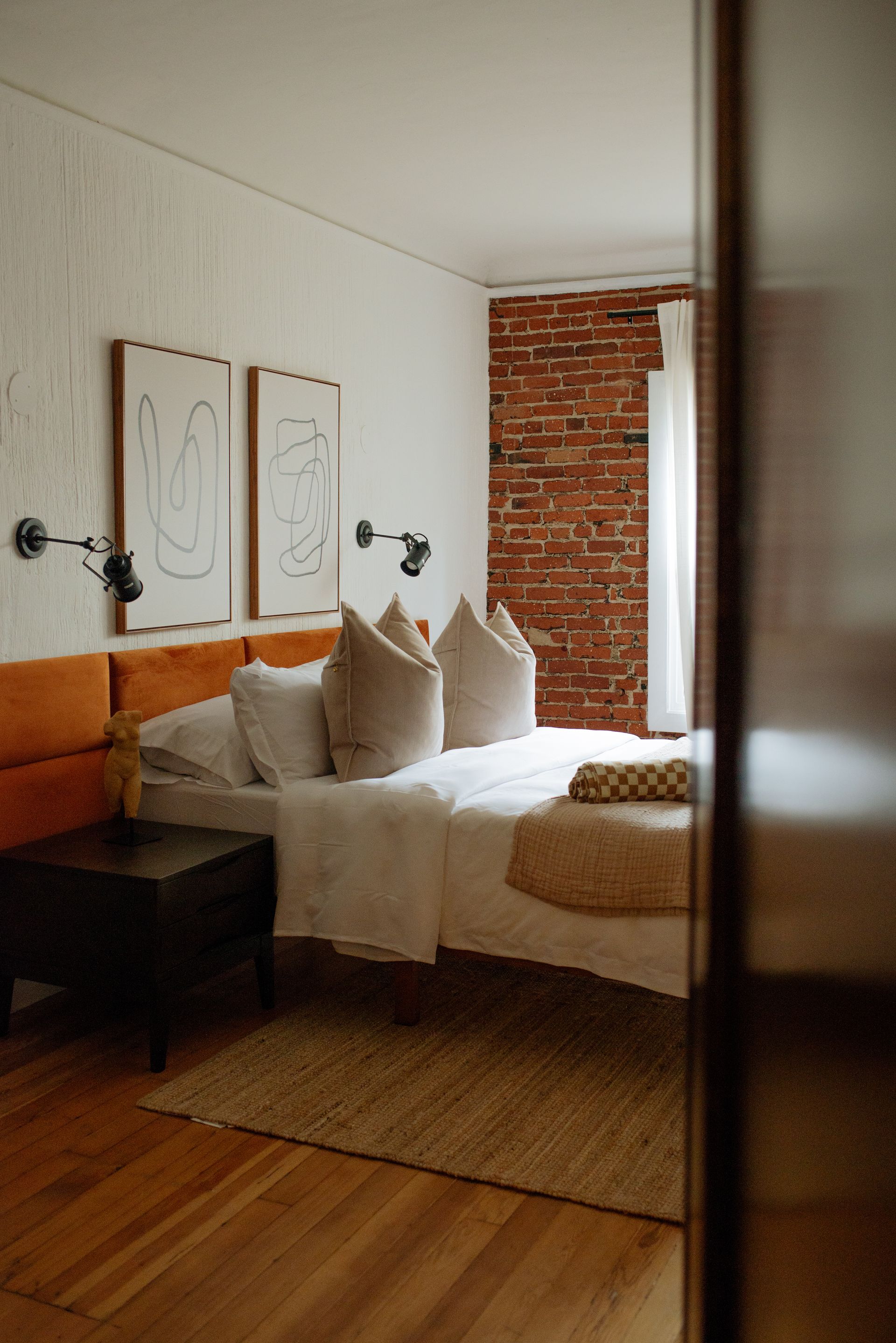 Bedroom with bed, orange headboard, brick wall, artwork, and jute rug.