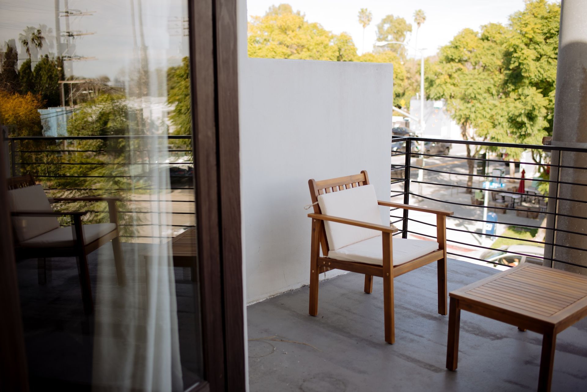 Balcony with wooden chair and table, white wall, metal railing, view of trees and city.