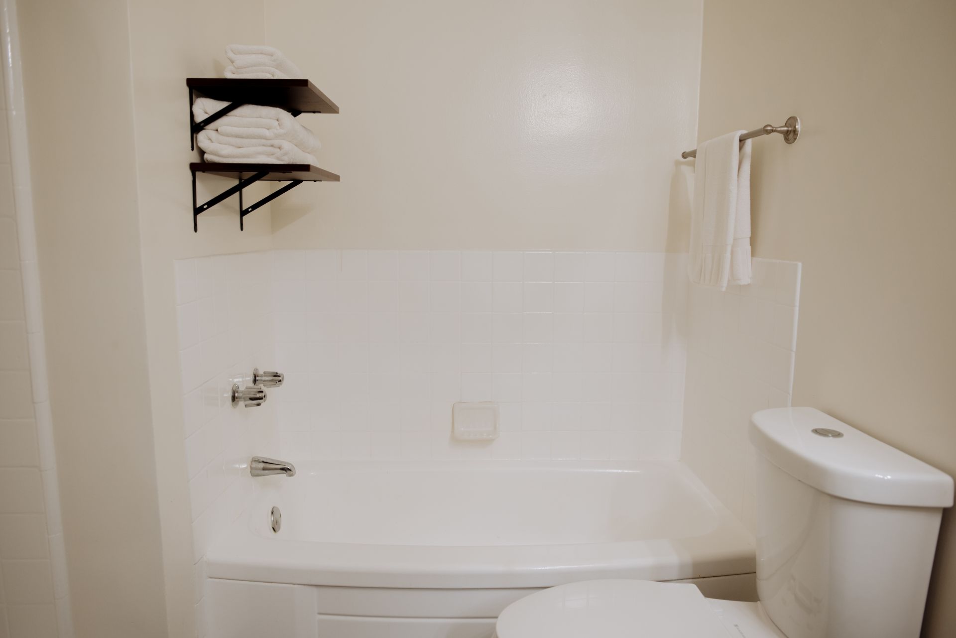 White bathroom with a tub, toilet, towels, and a shelf with folded towels.