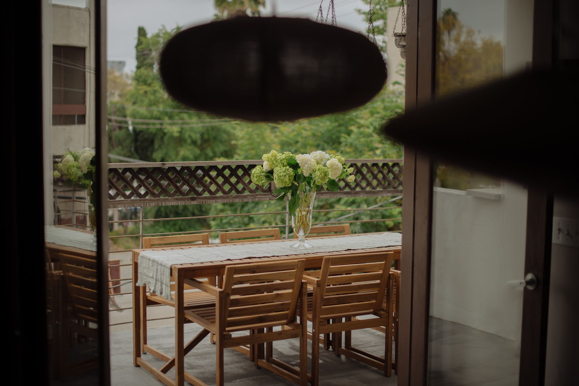 Wooden outdoor dining set with flowers, viewed through sliding glass doors.